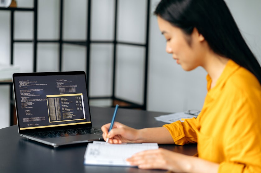 Woman taking notes with code on an open laptop next to her